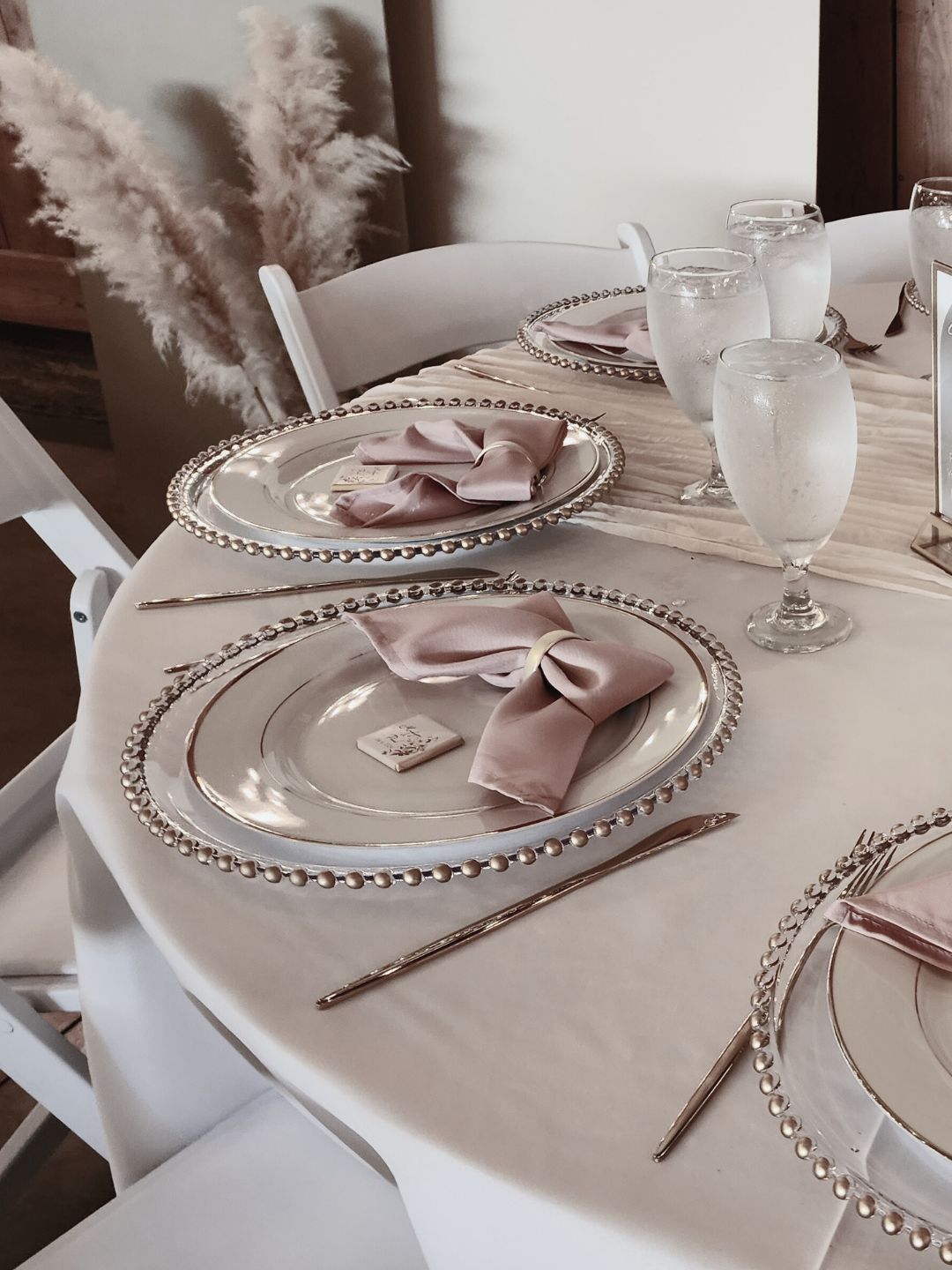 Closeup of reception tables featuring chargers with gold beads, white plates with gold rims gold flateware blush napkin folded into a bow with gold napkin ring and a square personalize chocolate favor