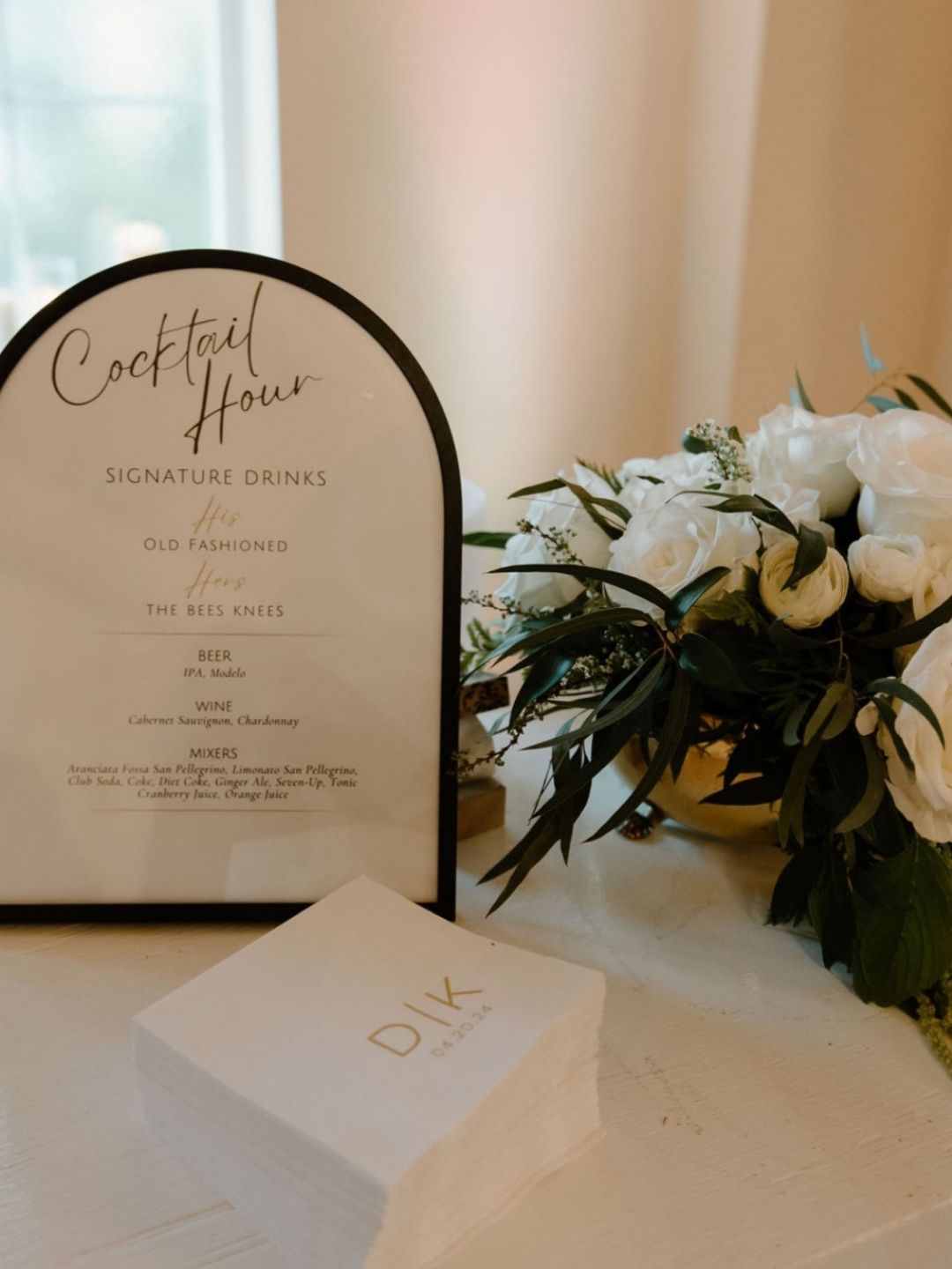 Cocktail Hour menu in a modern black arch frame with gold monogramed cocktail napkins and green and white floral arrangement in a round gold vase. 