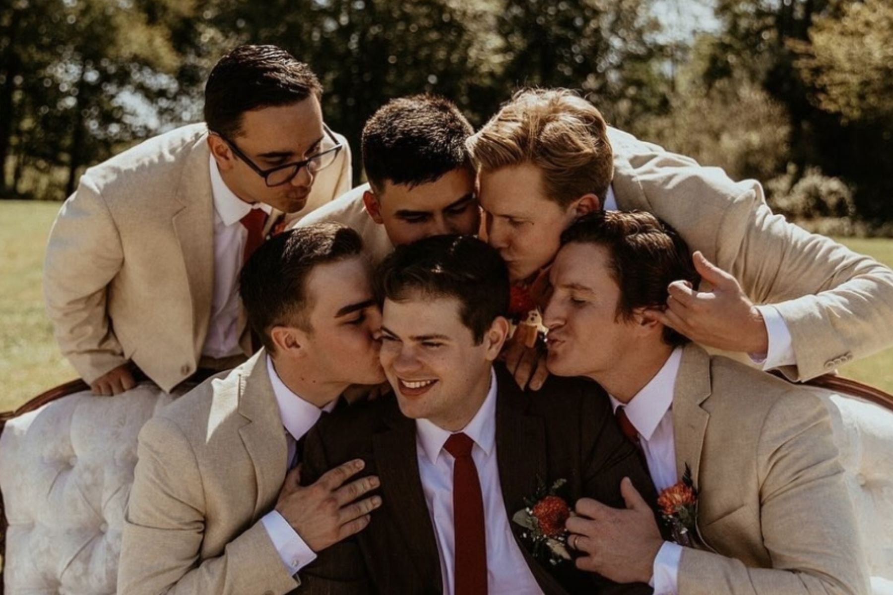 Smiling groom sitting outdoors at the wedding reception lounge sitting on a white vintage sofa surrounded by his groomsmen playfully leaning in to kiss on the head 