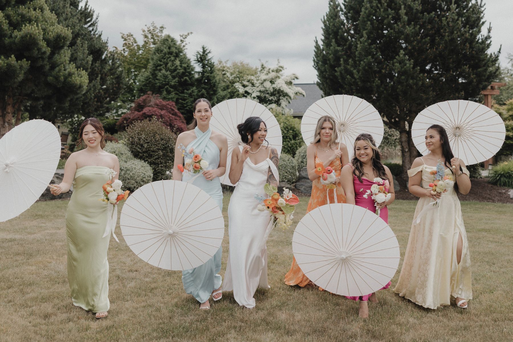 Bride and bridemaids walking and laughing while holding white paper parasols and citrus colored bouquets