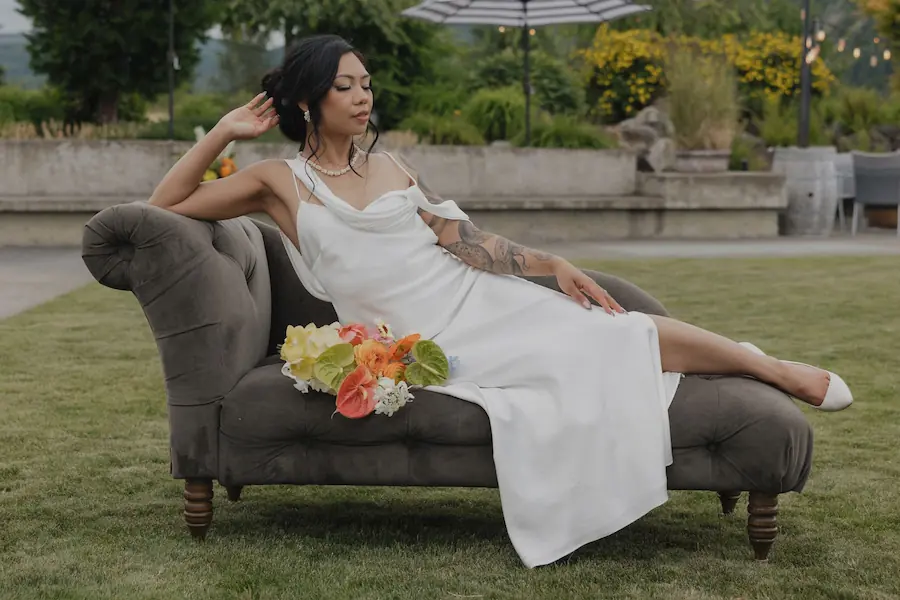 Bride in a white gown reclining on a vintage charcoal velvet settee on an outdoor estate lawn