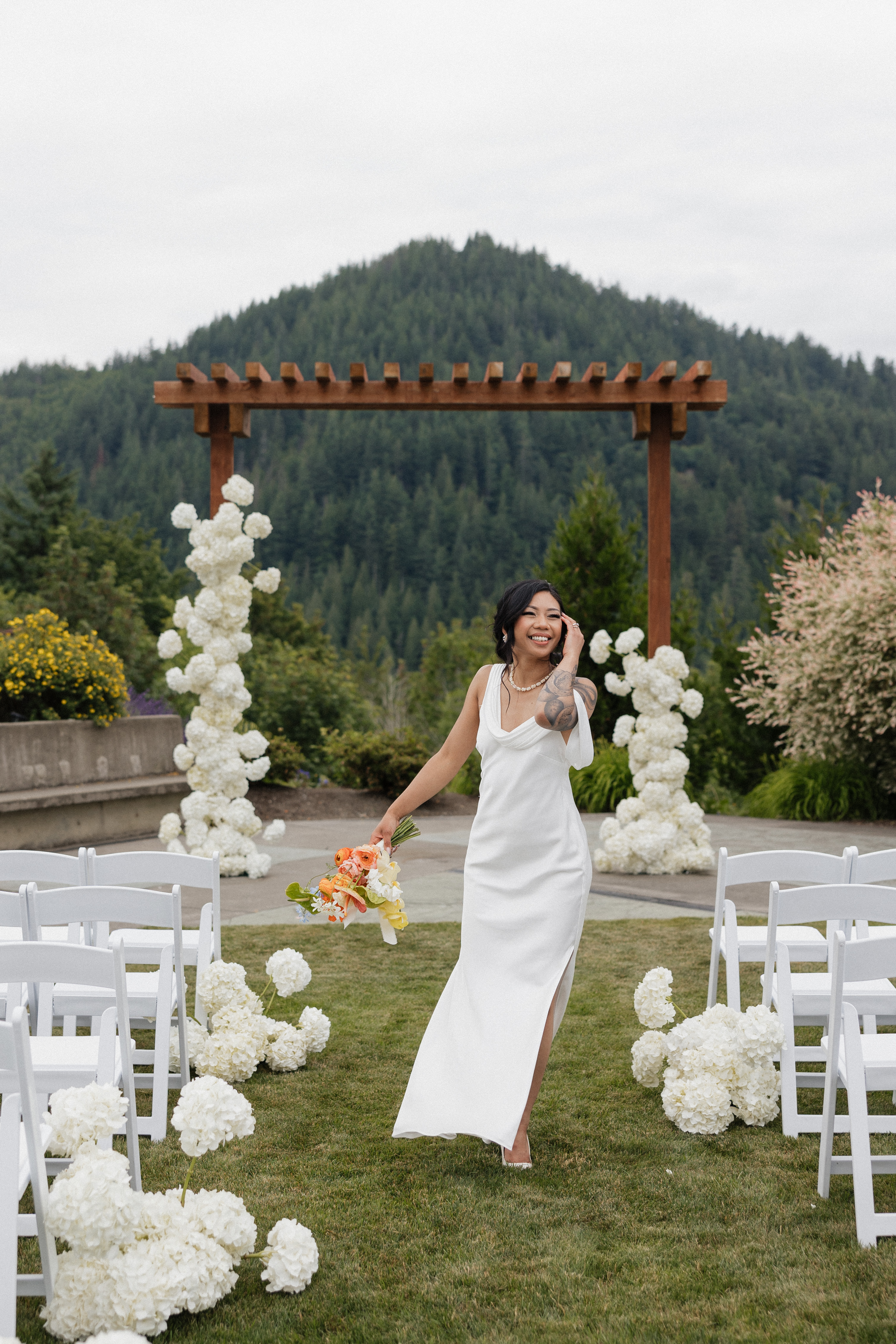 Modern bride in the PNW in her ceremony aisle with white chairs, white hydrangeas and wooden arch set against lust green pine mountain