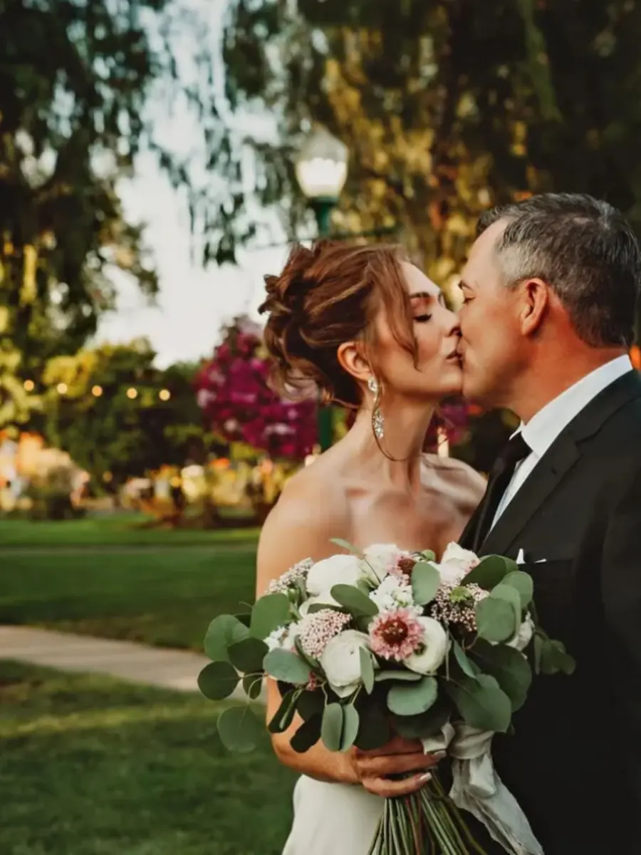 Bride holding a floral bouquet receiving a kiss from the groom outdoor garden setting