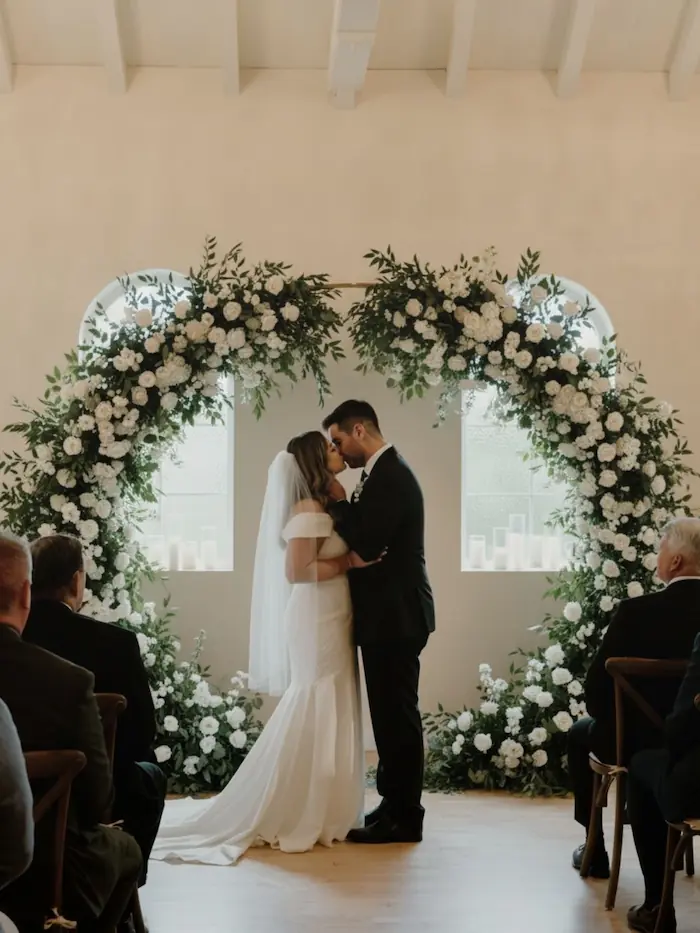Bride and groom kissing during a ceremony framed by a large circular white floral arch