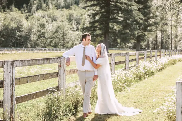 bride and groom by horse pasture with white wild flowers and evergreen trees