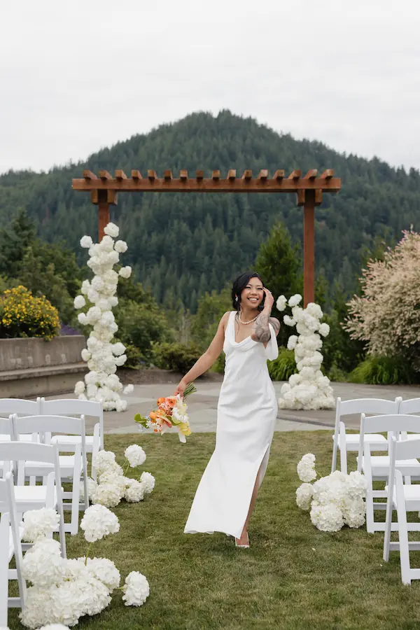 Modern bride in the PNW in her ceremony aisle with white chairs, white hydrangeas and wooden arch set against lust green pine mountain