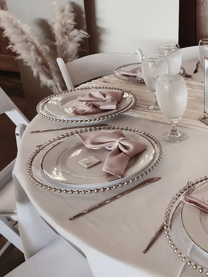 Closeup of reception tables featuring chargers with gold beads, white plates with gold rims gold flateware blush napkin folded into a bow with gold napkin ring and a square personalize chocolate favor