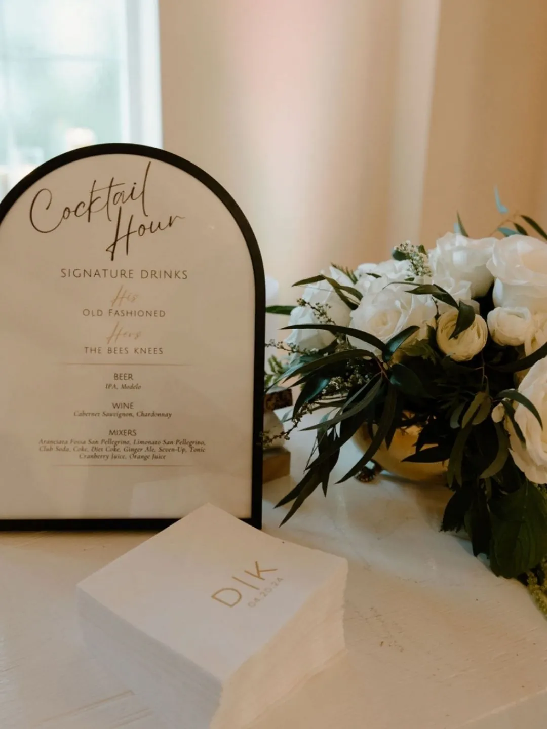 Cocktail Hour menu in a modern black arch frame with gold monogramed cocktail napkins and green and white floral arrangement in a round gold vase.
