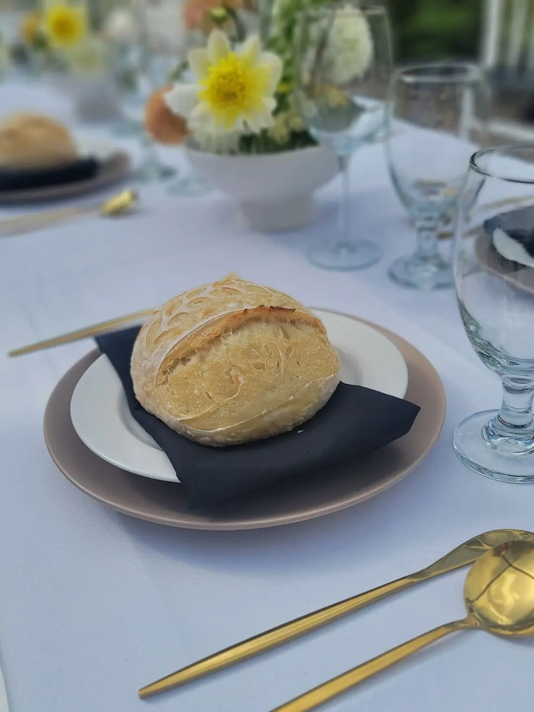 Close up of Modern minimalism tablesetting featuring stone dinnerware, black linen napkin topped with a personal sourdough boule from local artisan baker, gold flatware, clear glassware and yellow and peach ikebana floral arangements