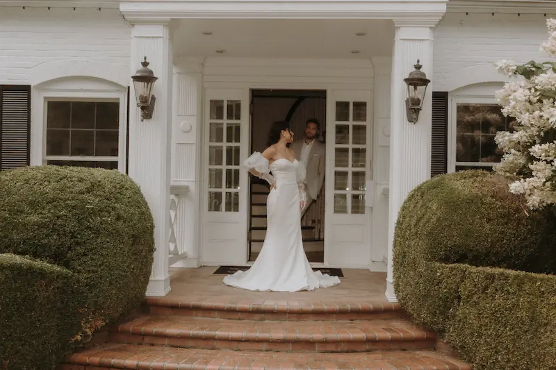 Bride in a white wedding gown standing on a stone pathway in front of a historic white estate