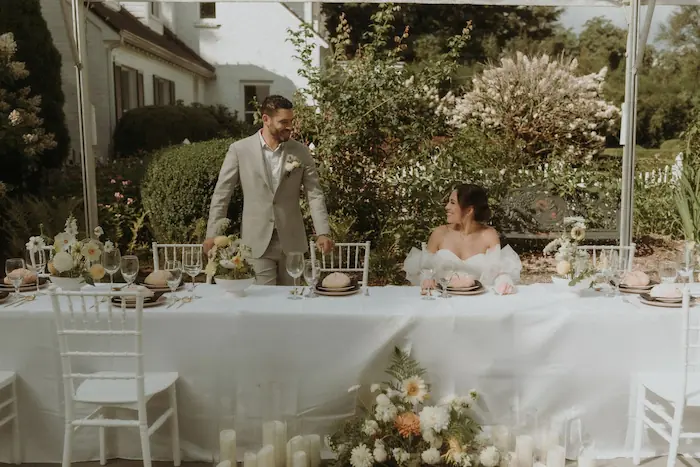 Bride and groom at an outdoor wedding reception table with white wooden chairs and lush greenery backdrop