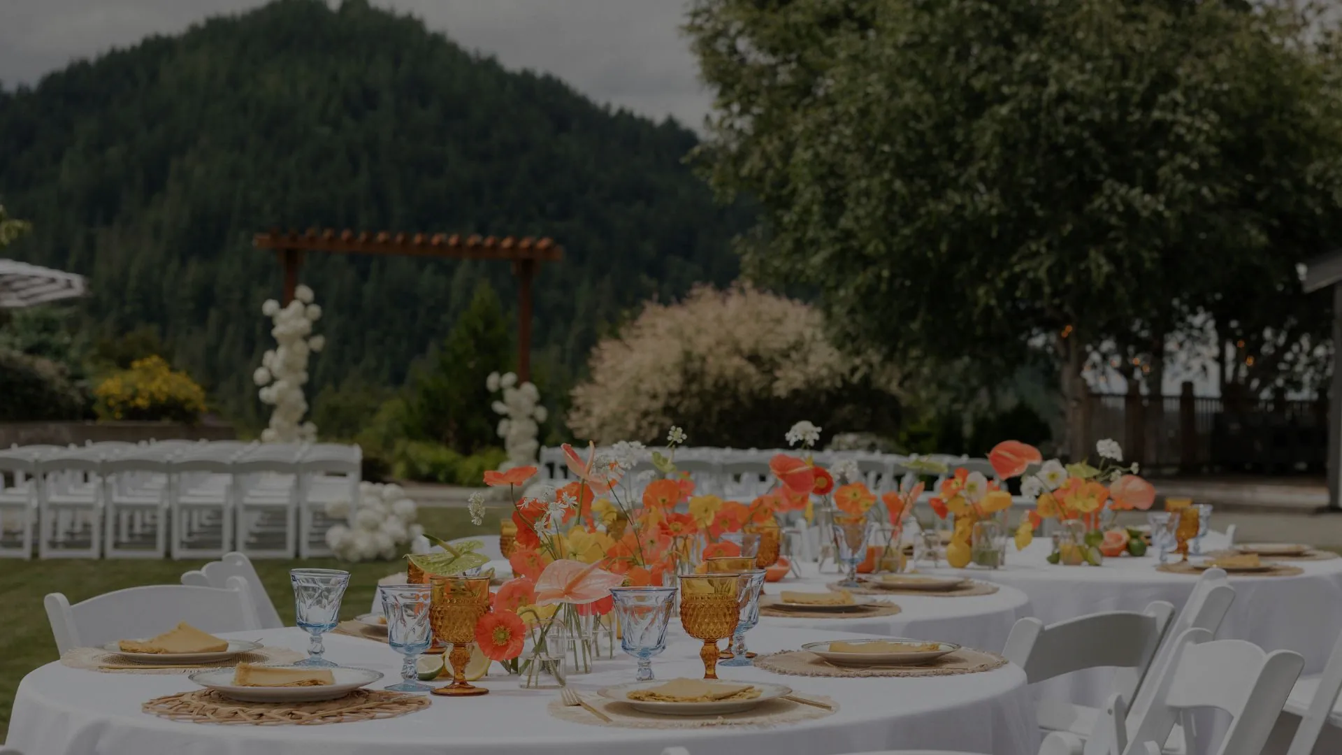 Outdoor wedding reception dining tables with white linens, floral centerpieces, and wooden cross-back chairs in a garden setting