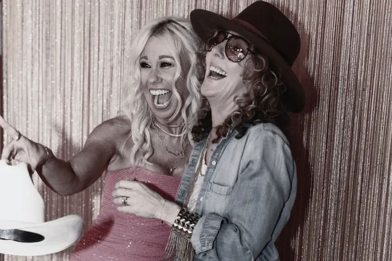 Two women laughing together in a photo booth wearing western outfits and cowboy hats
