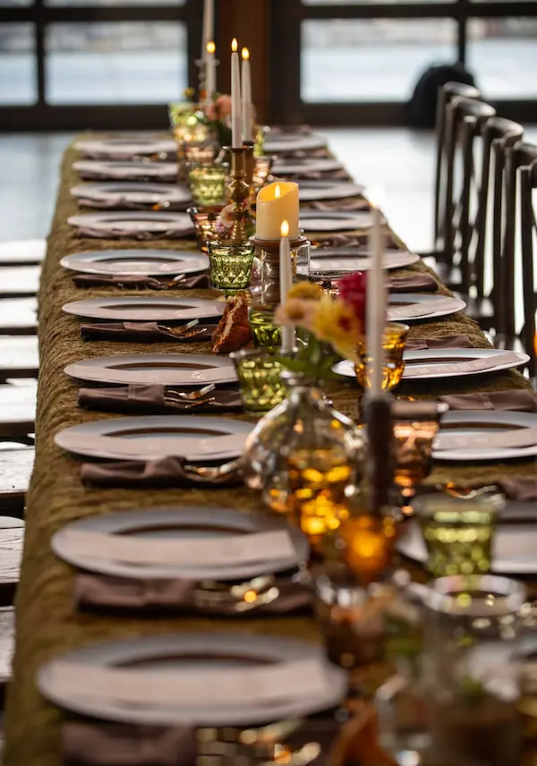 Long indoor reception table styled with a greenery garland runner, flickering taper candles, and wooden cross-back chairs