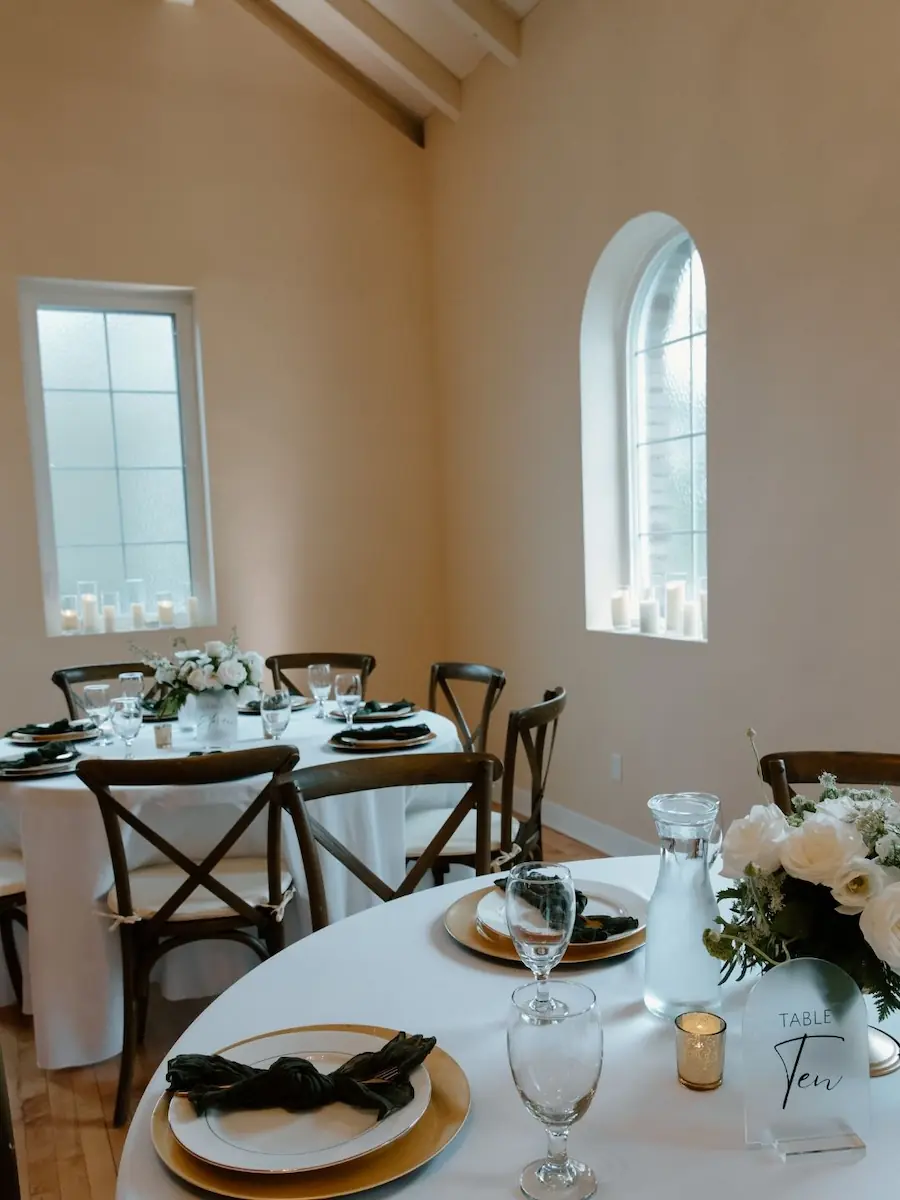 Indoor reception venue featuring large arched windows, round dining tables, and wooden cross-back chairs