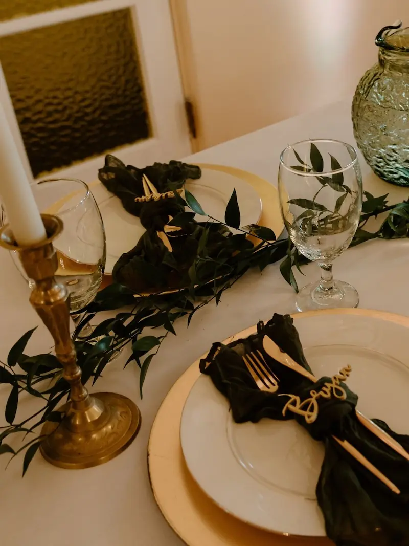 Table setting with gold flatware, olive green napkin, custom name tag, brass candlestick, and greenery