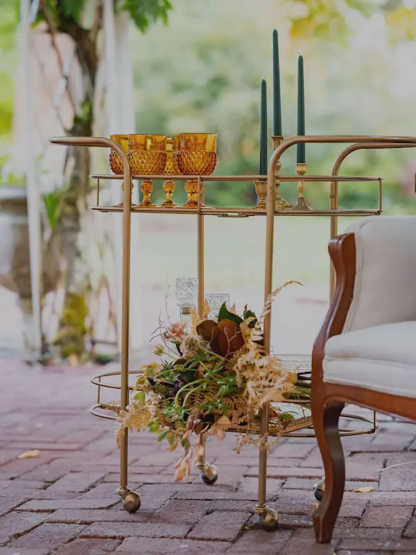 Close up of gold bar cart in garden lounge vignette with amber goblets, gold candlesticks fitted with dark green taper candles and a linen settee