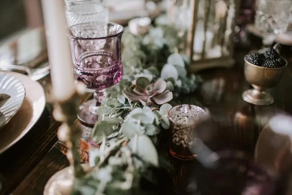 Closeup of wedding reception table decor featuring purple goblets brass candlesticks blackberries in brass bowls and succulent garland
