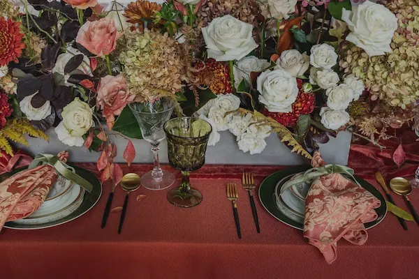 Table setting with red and pink floral centerpieces, dark green goblets, burgundy napkins, and brass cutlery