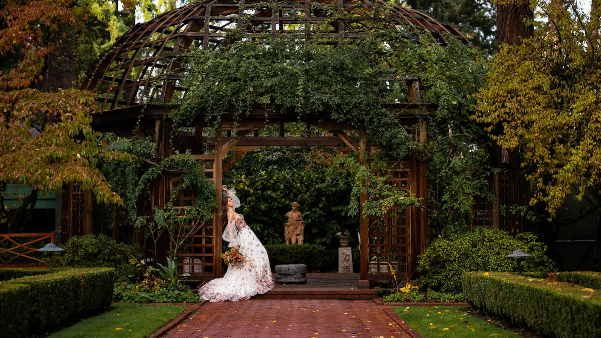 Bride in a white gown walking through a lush estate garden near an ornate iron gazebo