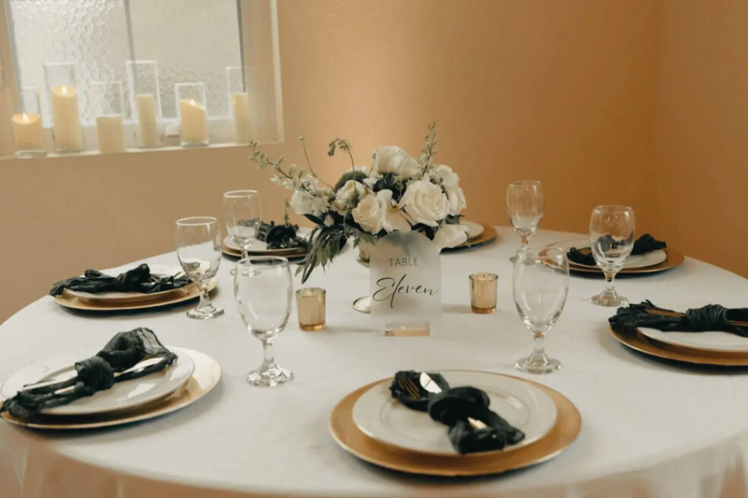 Minimalist Italian Wedding reception table set with gold chargers, ivory plates, olive green napkins, gold flatware, white and green floral centerpiece with candles flickering on the windowsill in the distance.