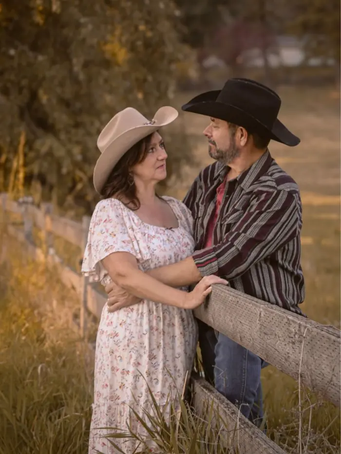 Couple in western outfits and cowboy hats standing together in a field at sunset